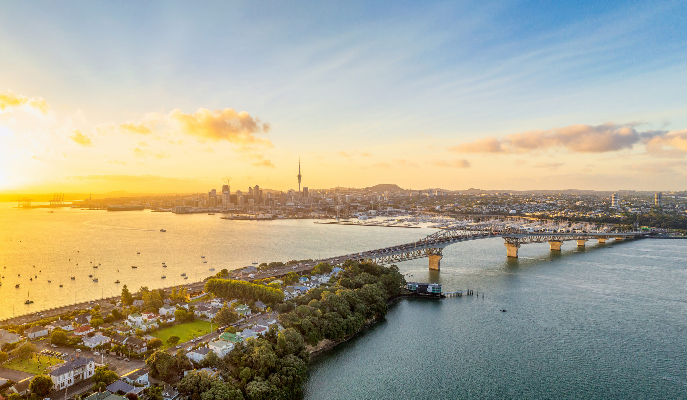 Aerial view of the Auckland skyline and Harbour Bridge at sunset during a family-trip, with boats on the golden water.