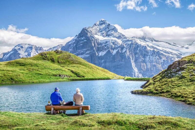 Two people sitting on a rustic bench looking toward a mountain lake and a massive snow-capped peak in Switzerland.