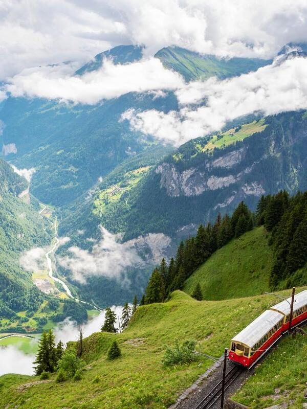 A red and white cogwheel train traveling up a lush green mountainside next to a deep valley filled with clouds.