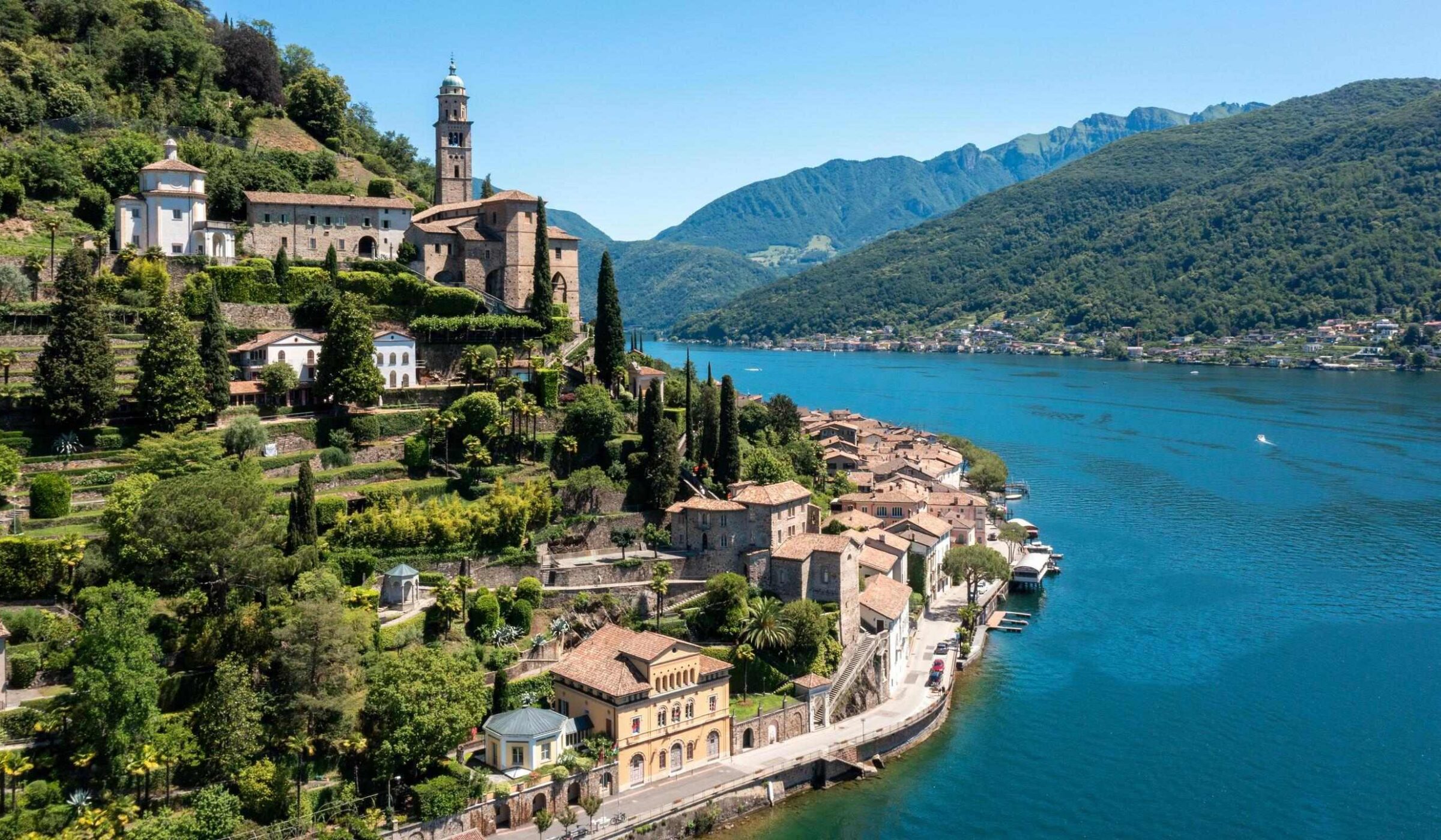 A hillside village featuring historic stone buildings and a church spire alongside a large blue lake framed by mountains.