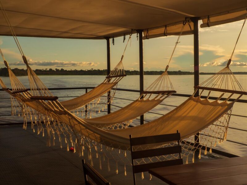 Three cream hammocks hang on a wooden boat deck overlooking a wide river at sunset on a latin america expedition cruise.