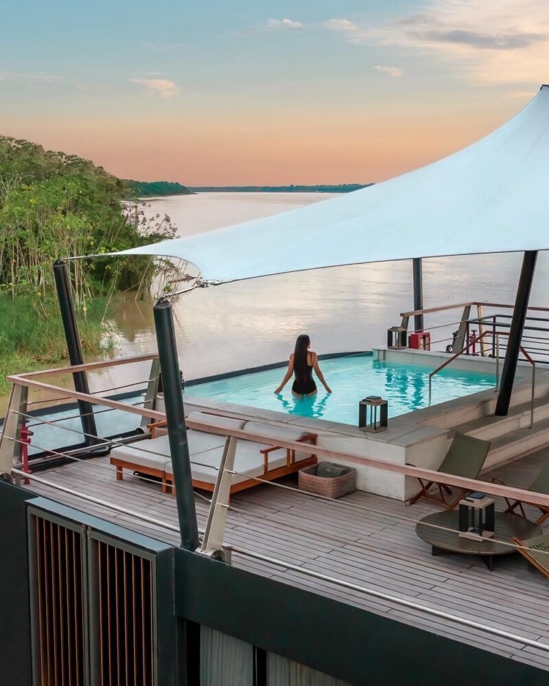 A woman stands in a shaded deck pool on a latin america expedition cruise, looking out at a wide river and green trees.