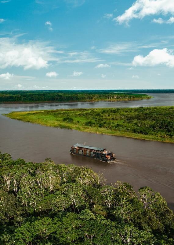 A modern latin america expedition cruise boat sails down a wide brown river surrounded by an expansive green rainforest.