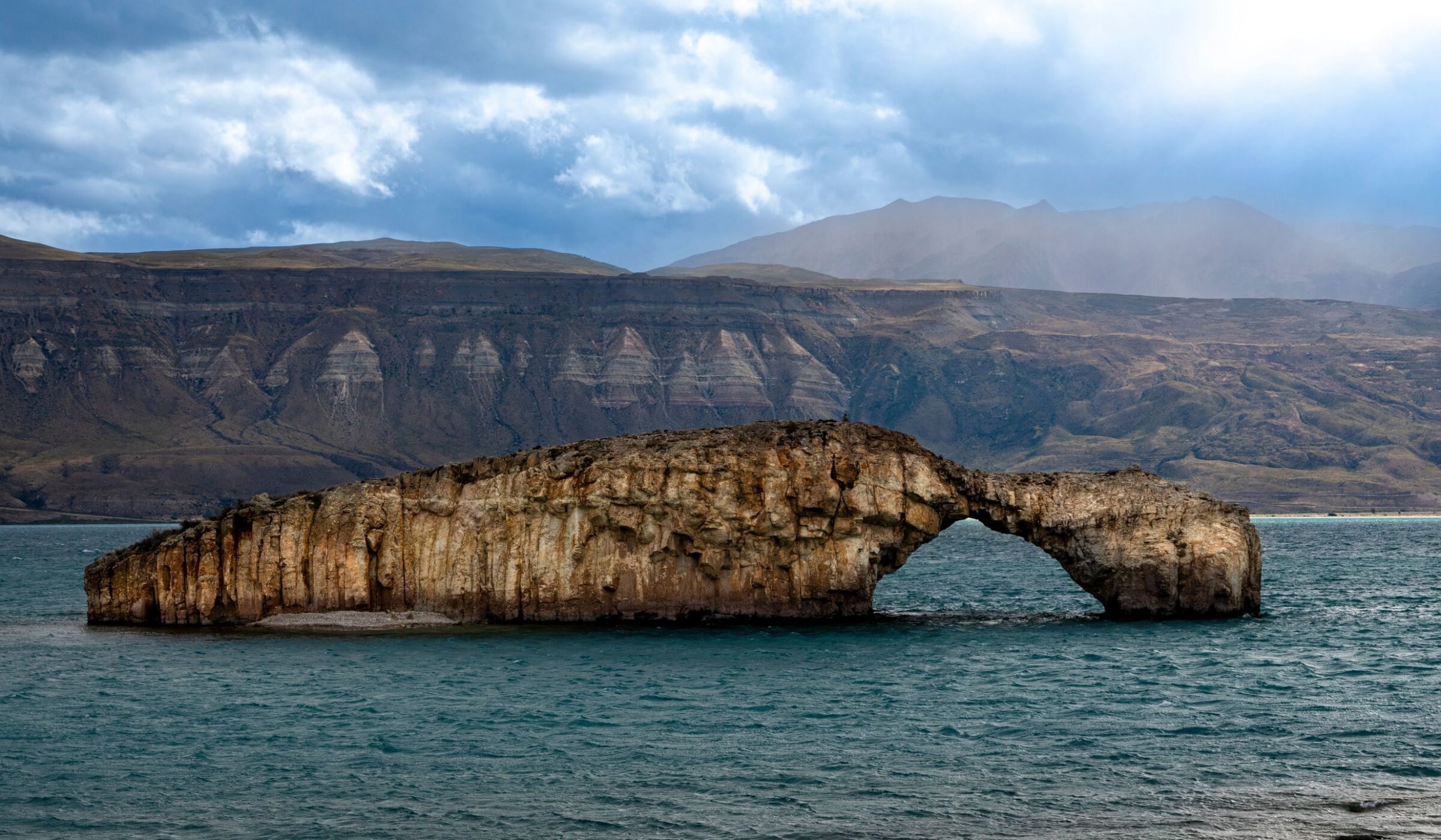 A large stone arch formation in a lake with mountains in the background in Posadas Argentina under a cloudy sky.