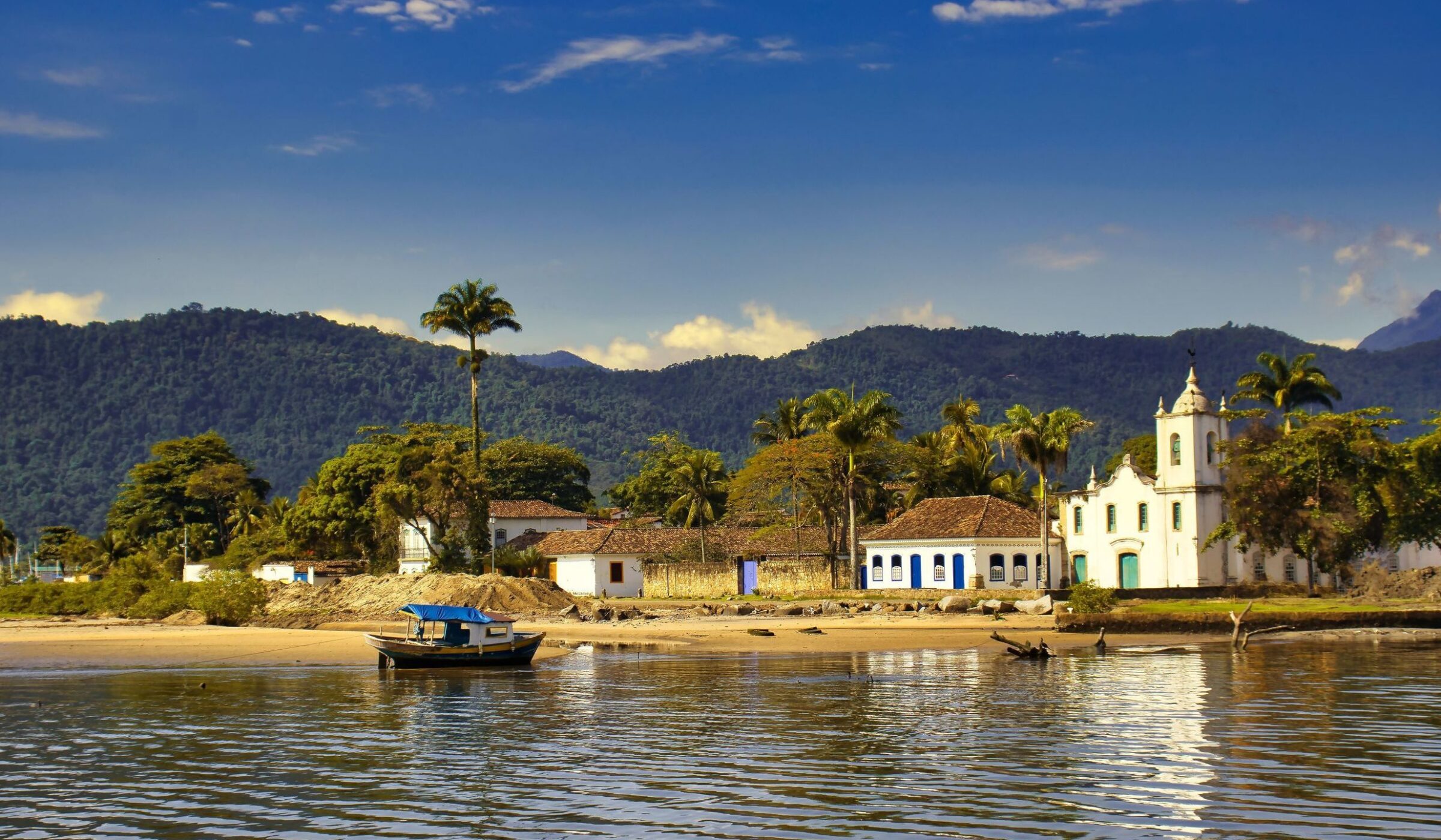 View from the water of white colonial buildings, a church, and a small boat on the shore in Paraty, Brazil.