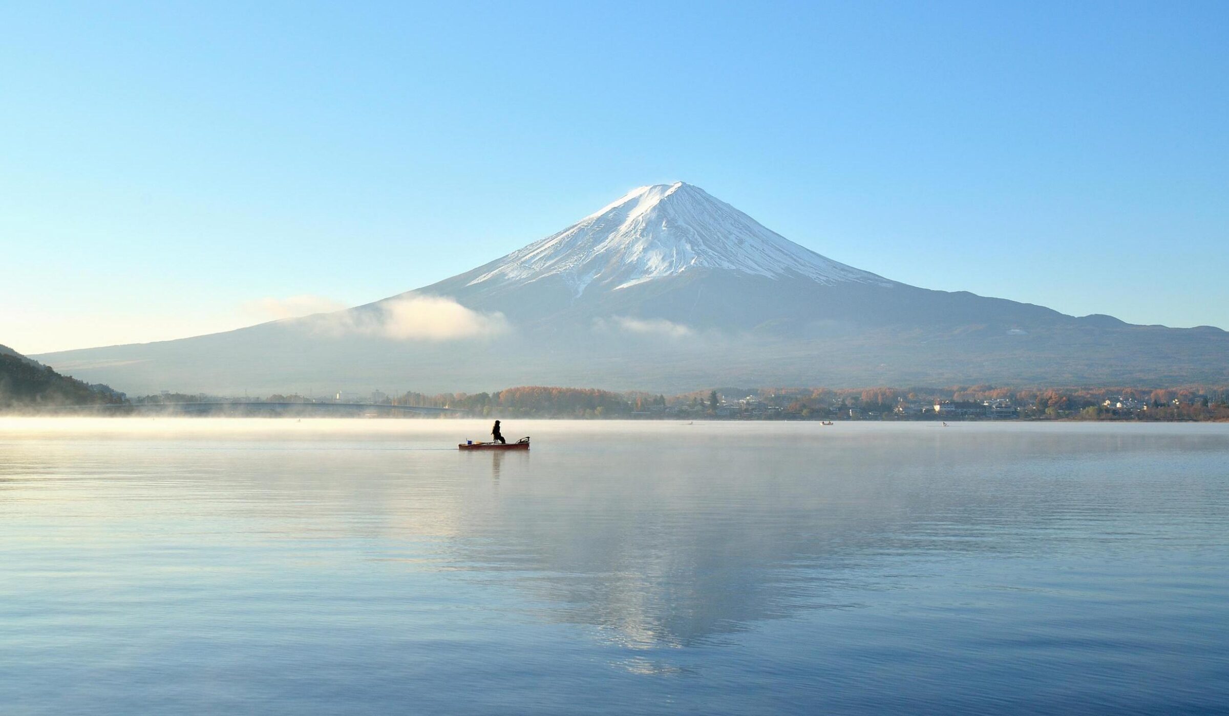 A small boat on a misty lake reflecting a large snow-capped mountain under a clear blue sky.