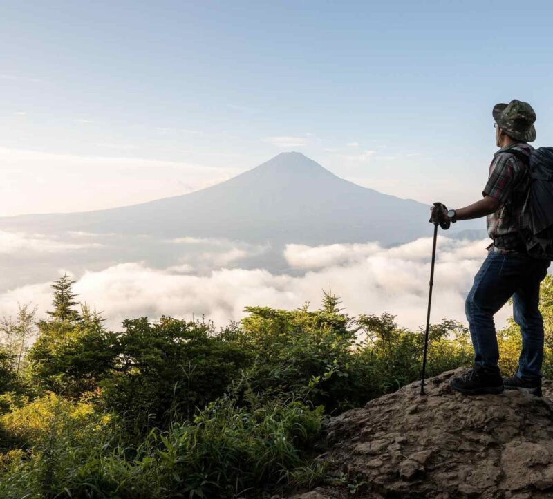 Hiker with a backpack and trekking pole standing on a rock looking at a distant mountain peak above clouds.
