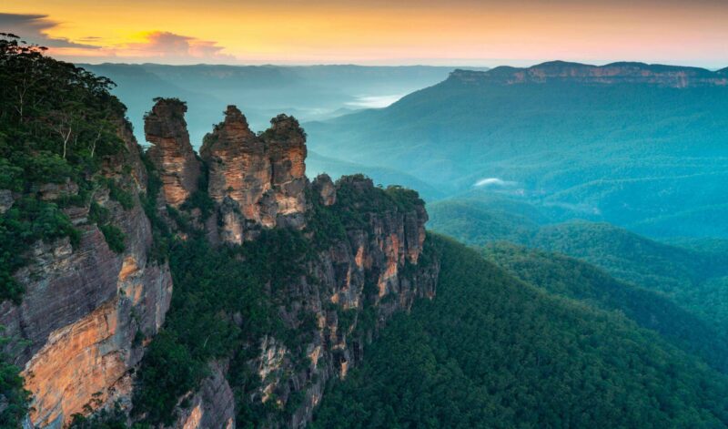 The Three Sisters rock formation rising over dense green forest and misty mountain valleys at sunrise.