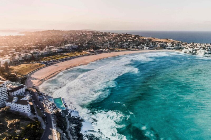 Aerial shot of Bondi Beach in Australia with turquoise ocean waves rolling onto a sandy shore next to coastal buildings.