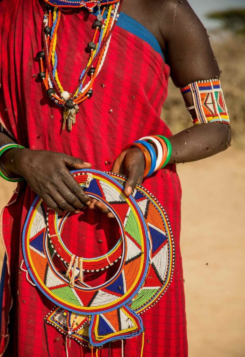 Midsection view of Maasai woman wearing a red garment holding intricate, colourful beaded collar necklaces.