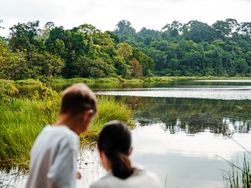 Two children look at a calm lake and dense green forest from the grassy edge of Khao Yai National Park.