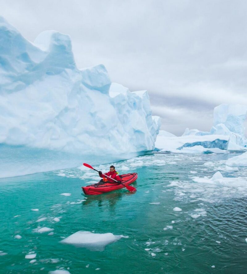 Person kayaking in a red kayak on bright turquoise water near massive icebergs during a polar safari.