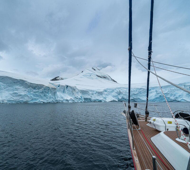 The bow of a sailing yacht approaches a colossal, towering glacier wall rising from the dark water during an exploratory polar safari.