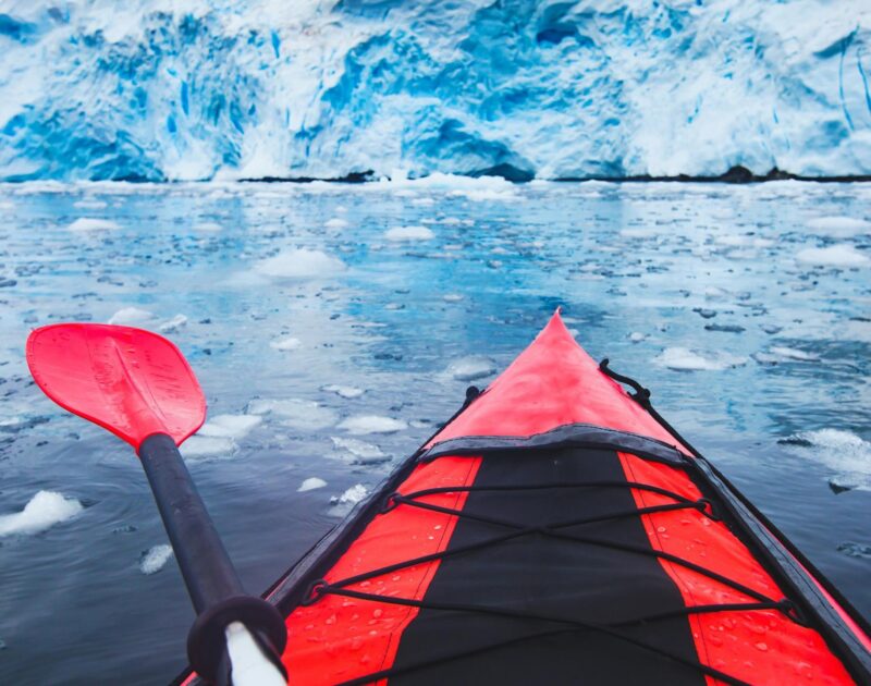 First-person view from a red kayak facing a massive blue ice wall in icy water on a polar safari.
