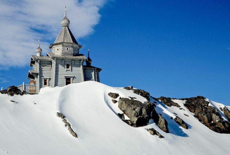 Wooden church with traditional onion domes perched on a snowy, rocky hill under a blue sky on a polar safari.