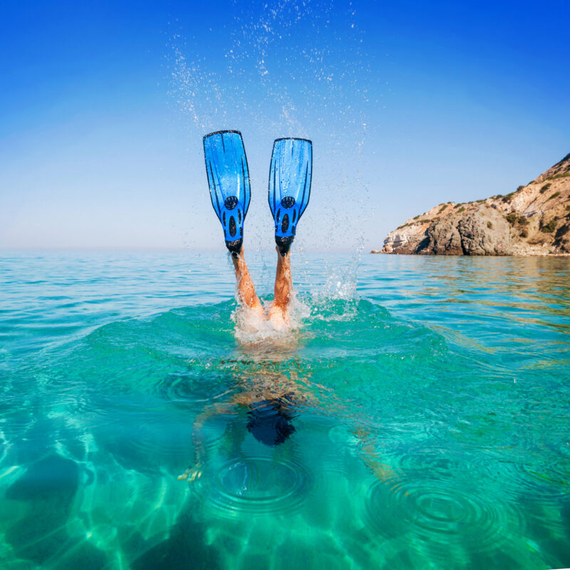 Blue snorkeling fins splashing above bright turquoise water as a person dives under, with a rocky coast behind.