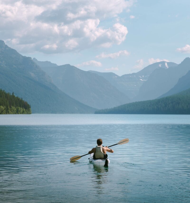 A man kayaks peacefully across a large body of water beneath some mountains