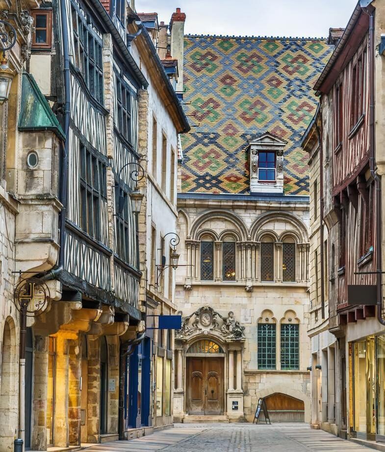 Narrow cobblestone street lined with half-timbered buildings leading to a stone facade with a brightly patterned tile roof.