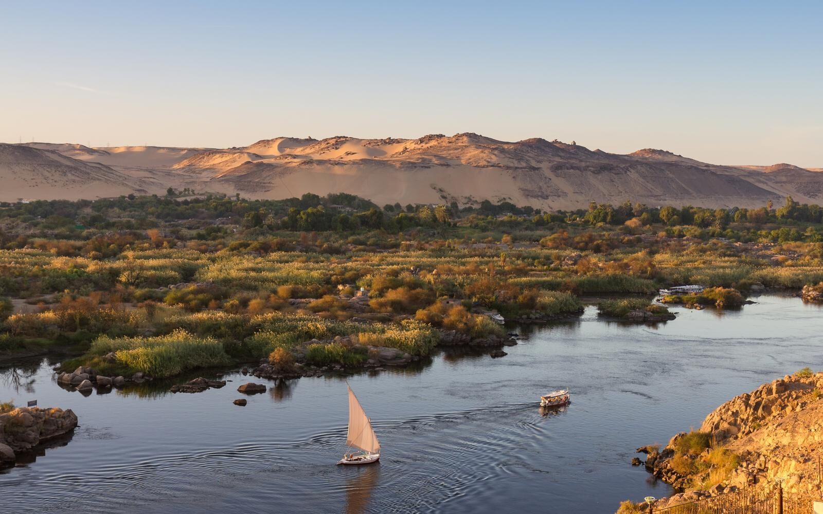 Sail along the Nile in a traditional felucca, passing lush riverbanks and golden desert hills around Aswan, Egypt.