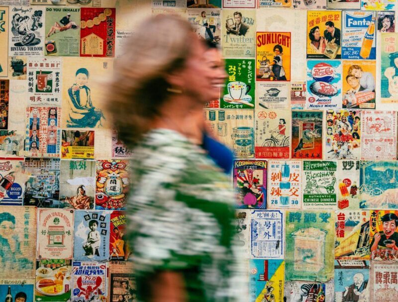 A mother and daughter on a trip exploring Bangkok's Chinatown walk past a wall adorned with vintage posters.