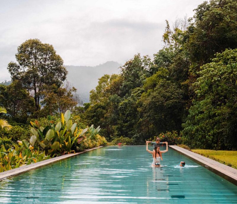 A family in a swimming pool at a resort in Thailand with Khao Sok's rolling hills in the background.
