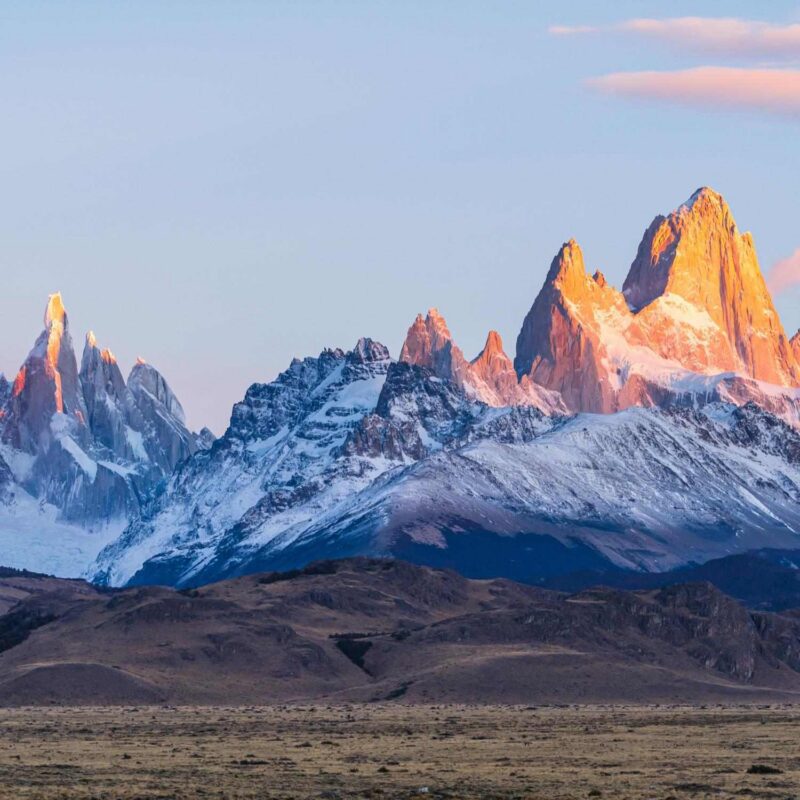 Jagged, snow-covered mountain peaks glowing orange under early morning light above flat plains.
