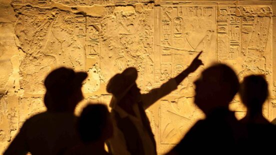 Silhouettes of tourists and a guide pointing at ancient Egyptian hieroglyphic stone carvings.