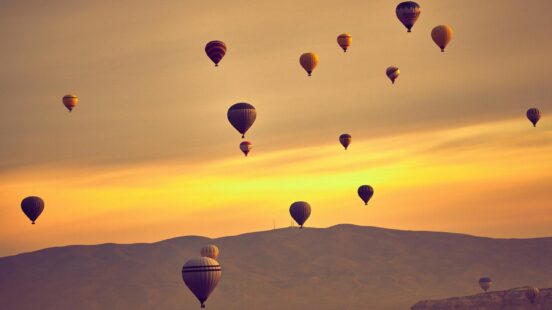Multiple hot air balloons floating in a warm golden sky above rolling mountain ridges.