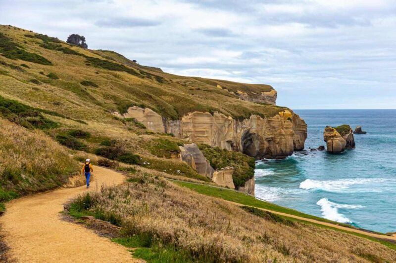 A lone hiker walking along a wide dirt cliffside trail next to ocean waves crashing against rocks.